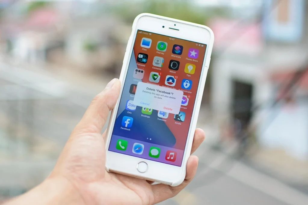 Osoba trzyma iPhone'a w swojej ręce. Na iPhone'ie wyświetla się komunikat z zapytaniem w języku angielskim, czy potwierdzić usunięcie aplikacji „Facebook”.