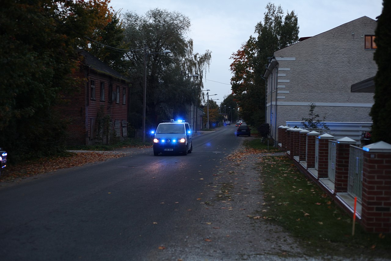 Zdjęcie przedstawia furgonetkę policyjną jadącą na sygnale po ulicy. Nieopodal znajdują się dwa domy – jeden wydaje się być pustostanem z cegieł i zabitymi oknami, a drugi ma kremowe wykończenie z prostymi dekoracjami.