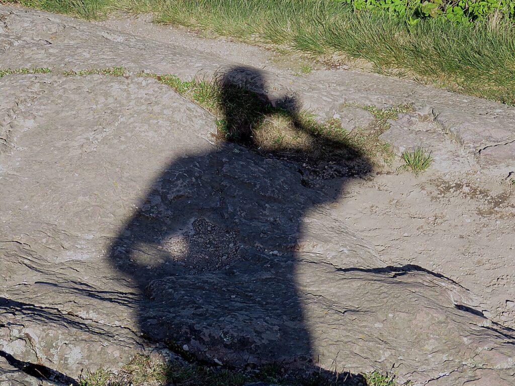 Zdjęcie przedstawia cień człowieka w tle najprawdopodobniej kamienistej, nierównej kładki oraz pasa zieleni.